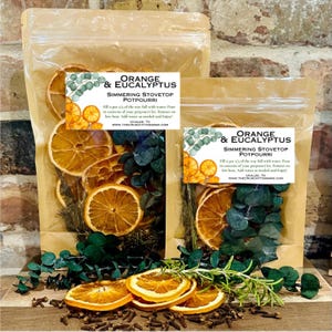 May include: Two clear bags filled with dried orange slices, eucalyptus, and spices. Each bag has a label that reads "Orange & Eucalyptus Simmering Stovetop Potpourri." The potpourri is arranged on a wooden surface.