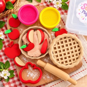 May include: A Play-Doh set featuring a tan pie crust, a wooden rolling pin, a red apple-shaped cookie cutter, and several red apple-shaped plastic pieces. The set includes two round Play-Doh containers in bright pink and yellow, plus a clear plastic storage box.