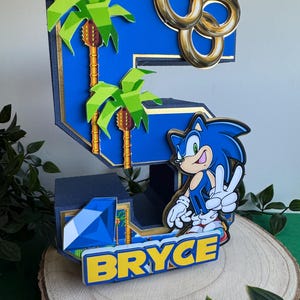 May include: A blue and yellow 3D cake topper shaped like the number 5, featuring Sonic the Hedgehog, palm trees, and gold rings. The topper also includes a blue gem and the name "BRYCE" in yellow letters.
