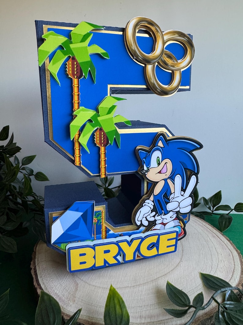 May include: A blue and yellow 3D cake topper shaped like the number 5, featuring Sonic the Hedgehog, palm trees, and gold rings. The topper also includes a blue gem and the name "BRYCE" in yellow letters.