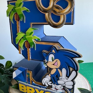 May include: A blue and gold 3D number five cake topper featuring Sonic the Hedgehog, palm trees, and gold rings. The topper has the name "Bryce" written on the base.