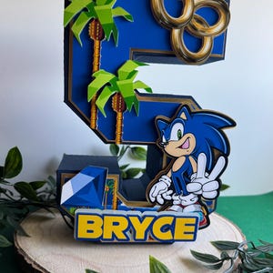 May include: A blue and gold 3D number five cake topper featuring Sonic the Hedgehog, palm trees, and gold rings. The topper also includes a blue gem and the name "Bryce" in yellow letters.