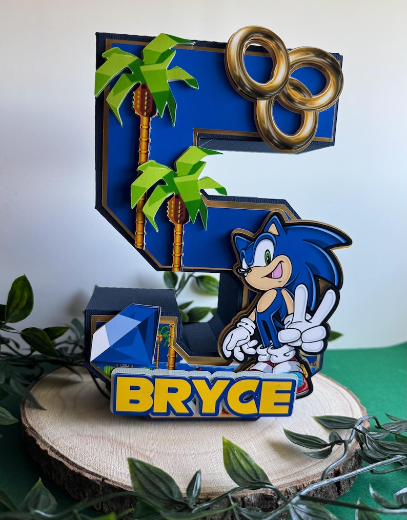 May include: A blue and gold 3D number five cake topper featuring Sonic the Hedgehog, palm trees, and gold rings. The topper also includes a blue gem and the name "Bryce" in yellow letters.