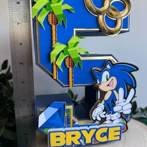 May include: A blue and gold 3D number five cake topper featuring a Sonic the Hedgehog character, palm trees, gold rings, and a blue diamond. The name "Bryce" is written in yellow letters on the base.