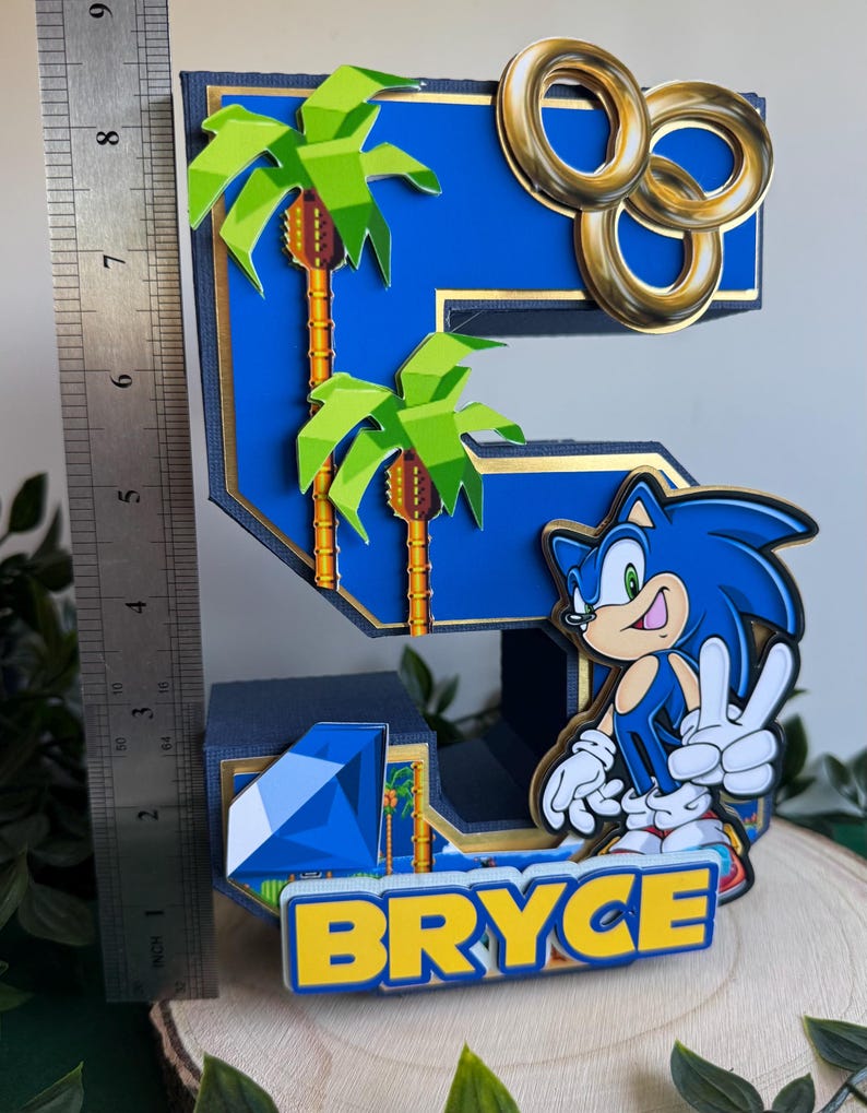 May include: A blue and gold 3D number five cake topper featuring a Sonic the Hedgehog character, palm trees, gold rings, and a blue diamond. The name "Bryce" is written in yellow letters on the base.