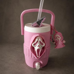 May include: A pink insulated beverage cooler featuring a white Ghostface mask emblem on the front, a white lid with a gray spout, a clear straw, and a matching pink Ghostface keychain attached to the side handle. The cooler has a white spigot.