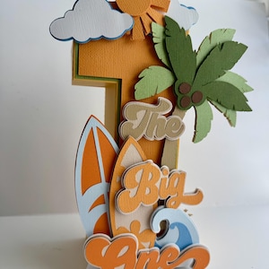 Peut inclure: Un topper de gâteau en papier 3D en forme de numéro un sur le thème de la plage. Le numéro un est orange et comporte une planche de surf, un palmier et un soleil. Le texte "The Big One" est écrit en lettres orange et brunes.