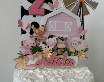 May include: A white cake with pink and gold farm-themed decorations. The cake is decorated with a pink barn, a windmill, a cow, a pig, a horse, a sheep, a chicken, butterflies, and the name "Julieta". The cake is decorated with a pink number 2.