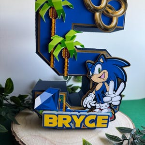 May include: A blue and gold Sonic the Hedgehog themed cake topper featuring the number 5, palm trees, gold rings, and a blue gem. The name "BRYCE" is written in yellow letters on the base.