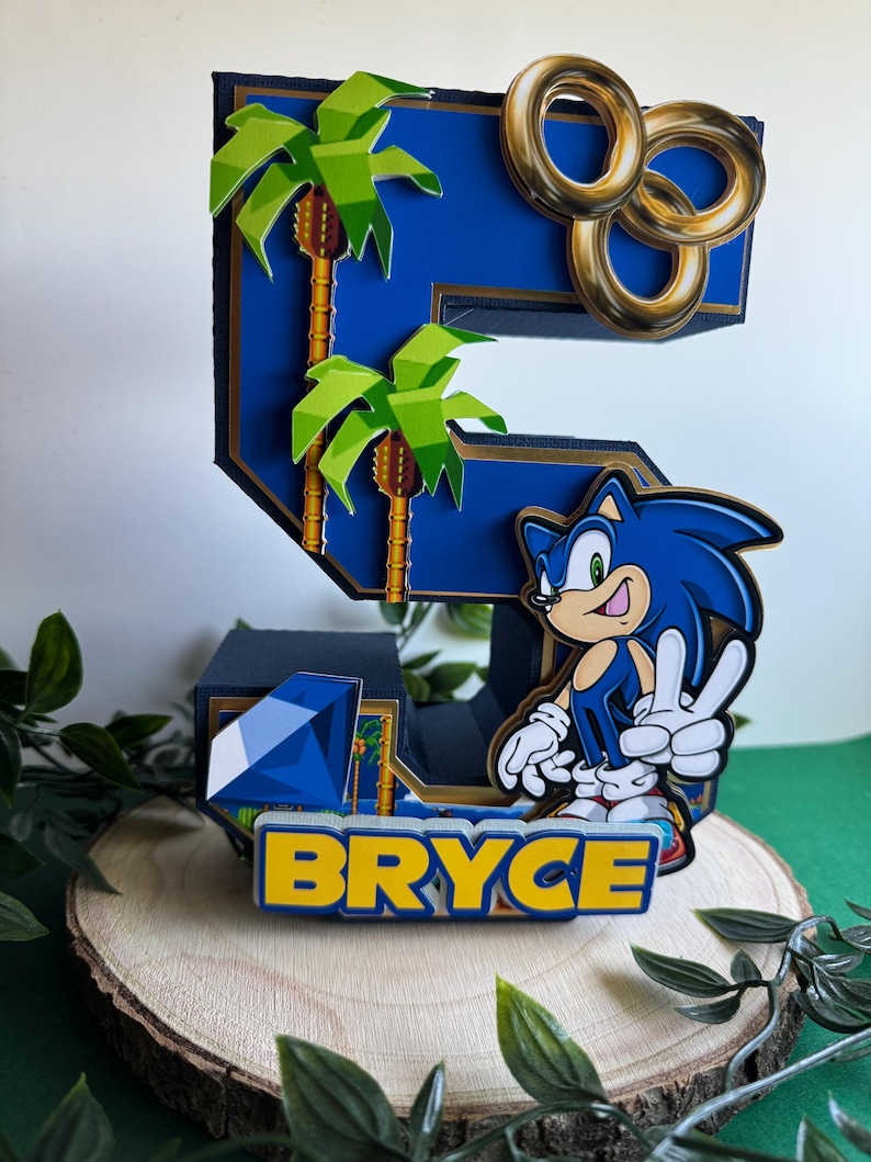 May include: A blue and gold Sonic the Hedgehog themed cake topper featuring the number 5, palm trees, gold rings, and a blue gem. The name "BRYCE" is written in yellow letters on the base.