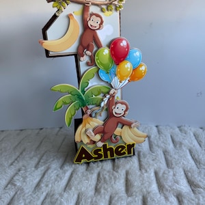 May include: A number one birthday decoration featuring a cartoon monkey holding balloons and bananas. The decoration is brown, yellow, green, red, and blue. The name "Asher" is printed on the base of the decoration.