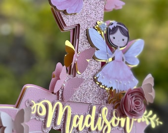 May include: A pink and gold glitter cake topper with a number one, a fairy, butterflies, and the name "Madison".