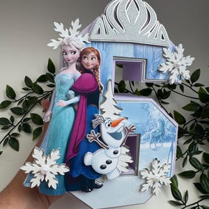 Könnte beinhalten: Eine dekorative Zahl sechs mit den Figuren Elsa, Anna und Olaf aus dem Film Frozen. Die Zahl ist hellblau mit einer silbernen Glitzerkrone und Schneeflocken-Akzenten. Die Figuren stehen vor einer Winterszene.