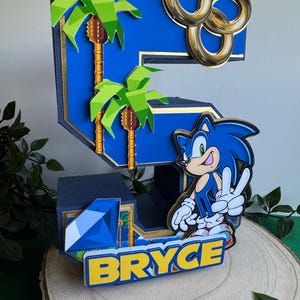 May include: A blue and gold 3D number five cake topper featuring a Sonic the Hedgehog character, palm trees, and gold rings. The name "Bryce" is written in yellow letters on a blue background.