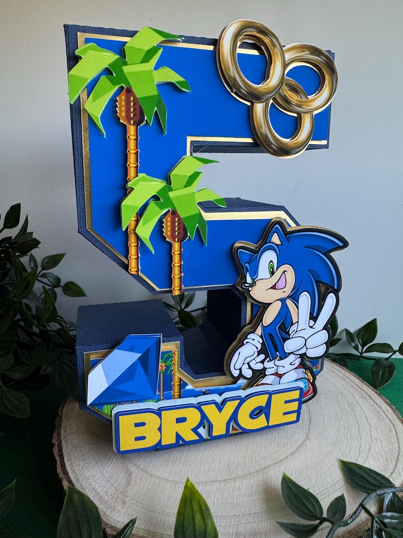 May include: A blue and gold 3D number five cake topper featuring a Sonic the Hedgehog character, palm trees, and gold rings. The name "Bryce" is written in yellow letters on a blue background.