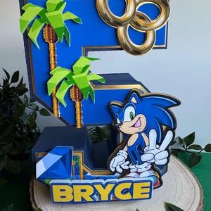 May include: A blue and gold 3D cake topper shaped like the number 5, featuring Sonic the Hedgehog, palm trees, and gold rings. The topper also includes a blue gem and the name "Bryce" in yellow letters.
