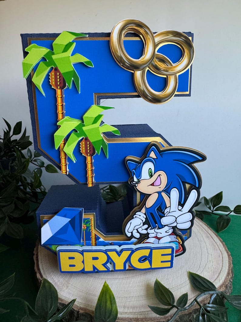 May include: A blue and gold 3D cake topper shaped like the number 5, featuring Sonic the Hedgehog, palm trees, and gold rings. The topper also includes a blue gem and the name "Bryce" in yellow letters.