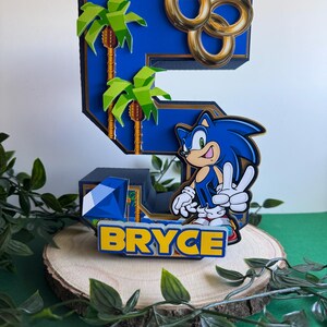 May include: A blue and gold 3D number five cake topper featuring a Sonic the Hedgehog character, palm trees, and gold rings. The topper also includes the name "BRYCE" in yellow letters.
