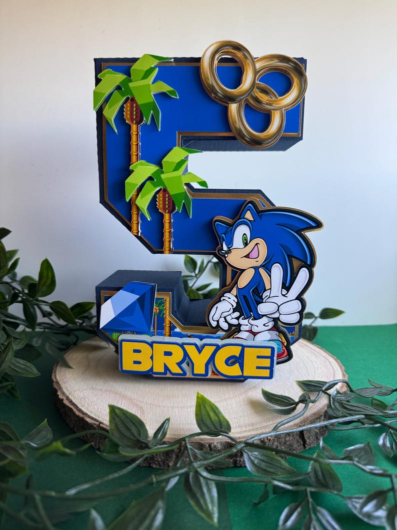 May include: A blue and gold 3D number five cake topper featuring a Sonic the Hedgehog character, palm trees, and gold rings. The topper also includes the name "BRYCE" in yellow letters.