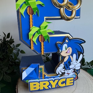 May include: A blue number five cake topper featuring Sonic the Hedgehog, palm trees, gold rings, and a blue gem. The name "BRYCE" is written in yellow letters at the bottom.