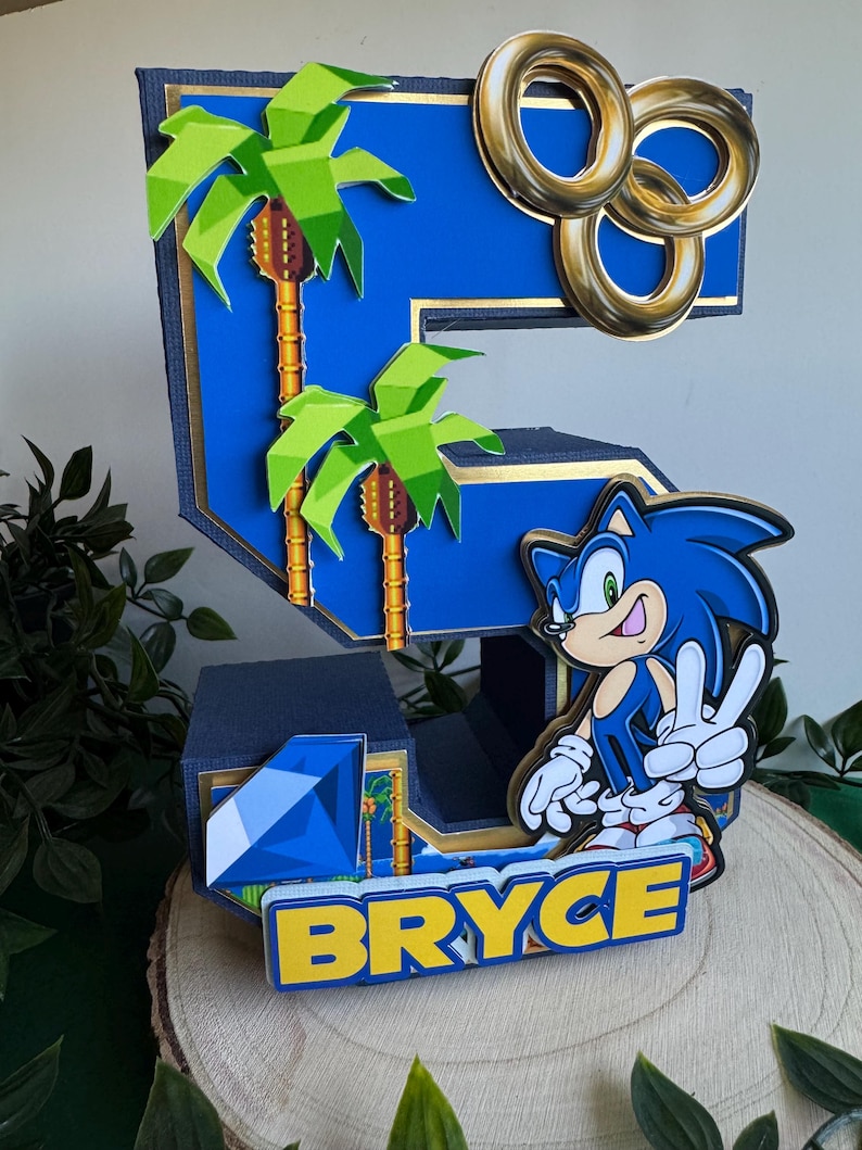 May include: A blue number five cake topper featuring Sonic the Hedgehog, palm trees, gold rings, and a blue gem. The name "BRYCE" is written in yellow letters at the bottom.