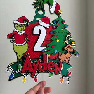 Pode incluir: Topo de bolo de Natal com tema Grinch com um Grinch verde usando um chapéu de Papai Noel, um enfeite vermelho com o número 2, uma árvore de Natal verde e o nome Ayden.