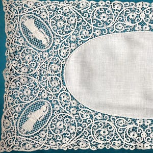 May include: Rectangular white lace doily with an oval center. The lace features a detailed floral and scroll design with four oval medallions. The doily is set against a teal background.