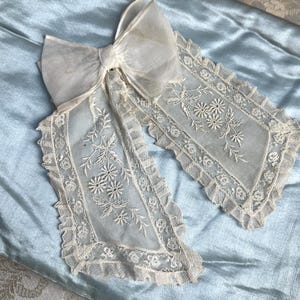 Puede incluir: Un cuello de encaje blanco vintage con un gran lazo y bordados florales. El cuello está sobre una tela de satén azul claro, con un fondo estampado floral. El encaje tiene un borde festoneado y delicadas flores bordadas.