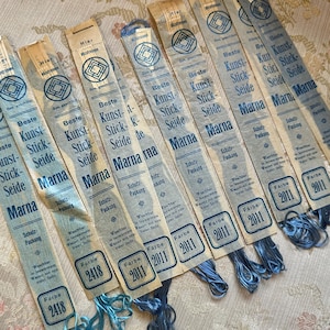 Könnte beinhalten: Neun einzelne Stränge Marna Stickgarn, jeweils umwickelt mit einem hellblauen Papieretikett mit Rautenlogo, dem Text Beste Kunst-Stick-Seide Marna und den Farbnummern 2418, 2011 oder 2015 mit blauen Fadenenden am unteren Ende.