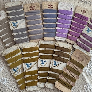 May include: A selection of vintage silk thread spools in shades of grey, blue, purple, and olive green. Each spool is labelled with French text and features decorative illustrations. The spools are arranged on a white lace surface.