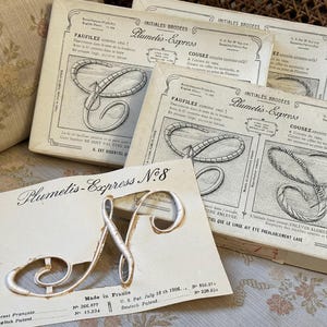 May include: Vintage Plumeitis-Express embroidery kits featuring the letter "N" and other letters. The kits include instructions and designs for embroidery. The packaging is aged with text in French and English.