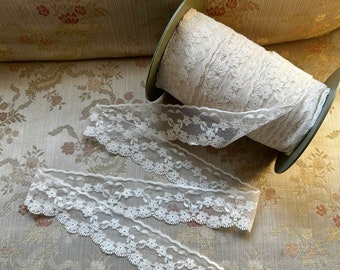 May include: A spool of cream-colored lace ribbon with a floral design, and two lengths of the same lace. The lace has a scalloped edge. The spool is on a floral patterned surface.