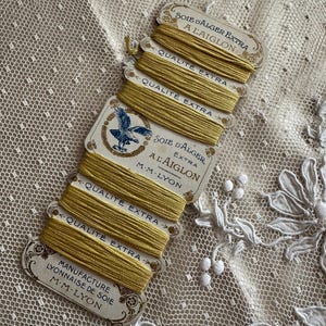 K&ouml;nnte beinhalten: Nahaufnahme von mehreren Spulen gelbem Seidenfaden, beschriftet mit "Soie d'Alger Extra A Laiglon" und "Qualit&eacute; Extra". Die Etiketten zeigen ein Adleremblem und stammen von "Manufacture Lyonnaise de Soie M.M. Lyon."