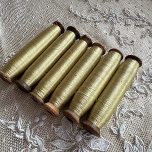 May include: Six spools of golden-yellow thread are arranged diagonally across a white lace surface. The thread is tightly wound on wooden spools with dark brown ends. The lace has a delicate floral pattern.