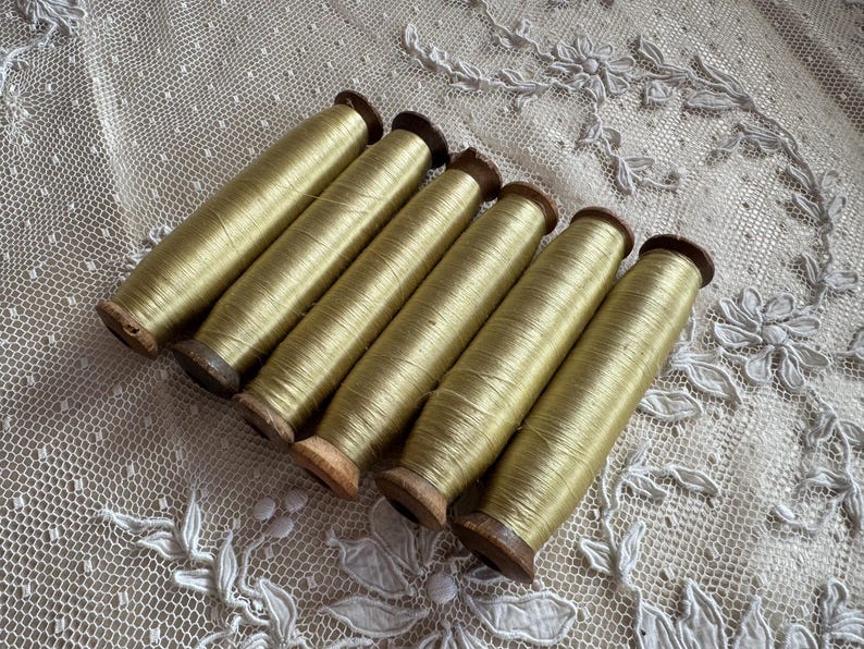 May include: Six spools of golden-yellow thread are arranged diagonally across a white lace surface. The thread is tightly wound on wooden spools with dark brown ends. The lace has a delicate floral pattern.