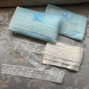Puede incluir: Dos paquetes de tela azul claro con ribete de encaje blanco, junto con dos rollos de encaje en crema y blanco. El encaje tiene intrincados patrones florales. Los artículos se muestran sobre una tela estampada floral.