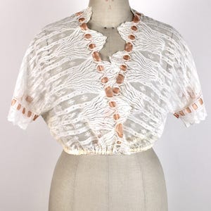 May include: A white lace top with short sleeves and a V-neckline. The top features a delicate pattern and is adorned with satin ribbon accents. The garment is displayed on a mannequin.