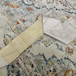 May include: A white fabric strap with a textured, patterned underside. The strap is folded in half and lying on a patterned rug.