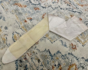 Puede incluir: Una correa de tela blanca con un lado inferior texturizado y estampado. La correa está doblada por la mitad y se encuentra sobre una alfombra estampada.