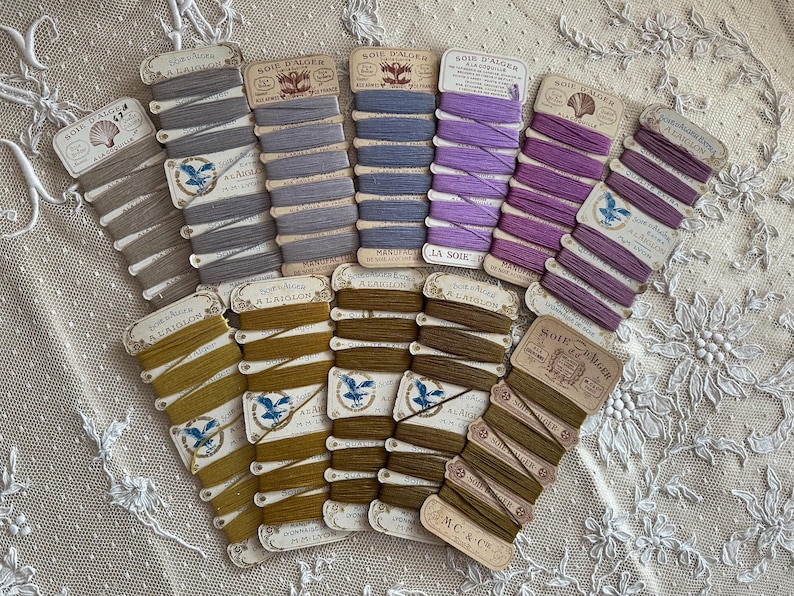May include: A selection of vintage silk thread spools in shades of grey, blue, purple, and olive green. Each spool is labelled with French text and features decorative illustrations. The spools are arranged on a white lace surface.