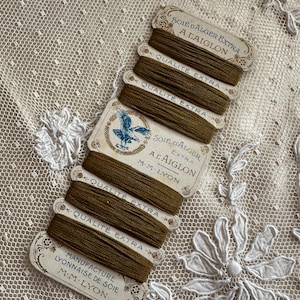 K&ouml;nnte beinhalten: Eine Sammlung von Vintage-Spulen aus olivgr&uuml;ner Seide mit dekorativen Etiketten. Die Etiketten tragen franz&ouml;sischen Text, darunter "Soie d'Alger Extra" und "Qualit&eacute; Extra", sowie ein blaues Adleremblem. Die Spulen sind auf einem wei&szlig;en Spitzengrund angeordnet.