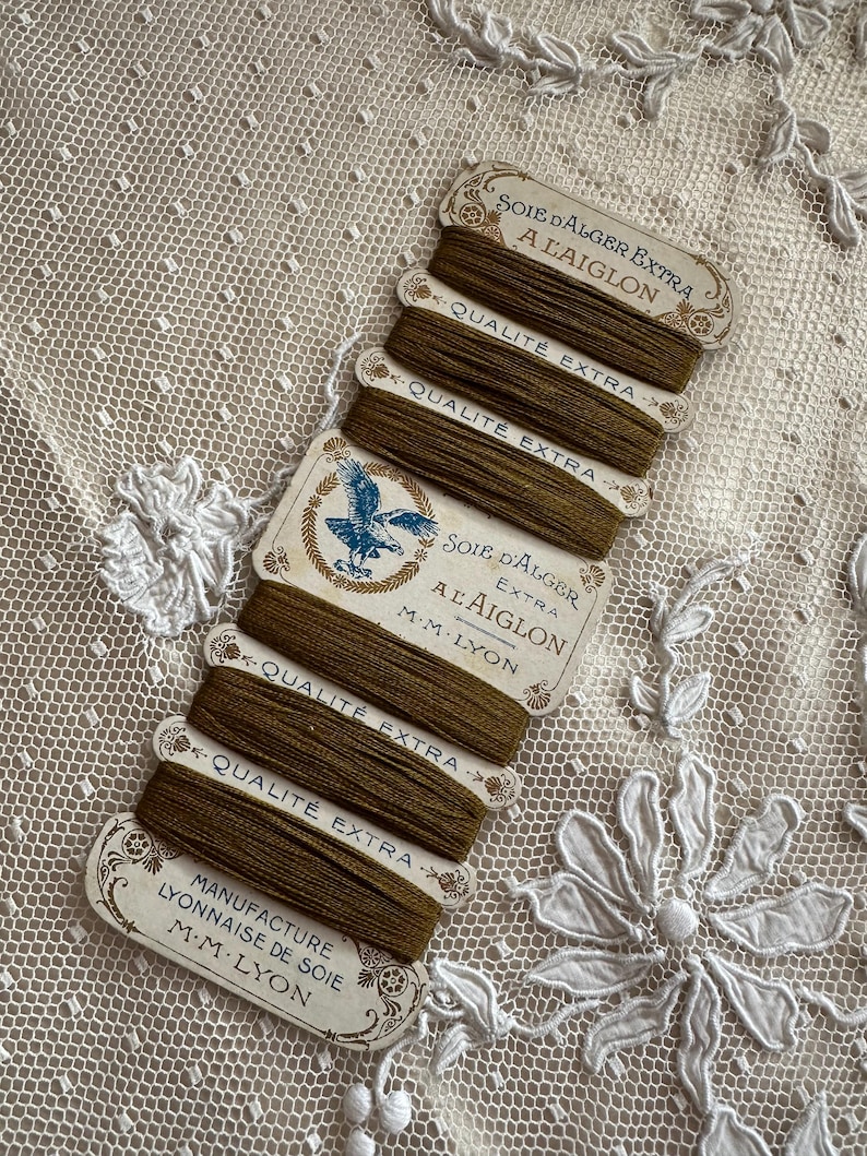 K&ouml;nnte beinhalten: Eine Sammlung von Vintage-Spulen aus olivgr&uuml;ner Seide mit dekorativen Etiketten. Die Etiketten tragen franz&ouml;sischen Text, darunter "Soie d'Alger Extra" und "Qualit&eacute; Extra", sowie ein blaues Adleremblem. Die Spulen sind auf einem wei&szlig;en Spitzengrund angeordnet.
