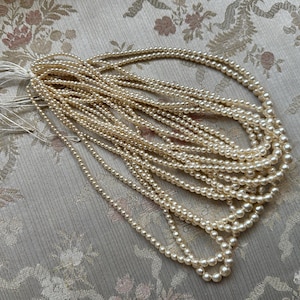 Puede incluir: Una colecci&oacute;n de collares de perlas de imitaci&oacute;n de color crema, colocados sobre una tela con estampado floral. Las perlas est&aacute;n ensartadas, creando m&uacute;ltiples hilos. Los collares son un accesorio cl&aacute;sico.
