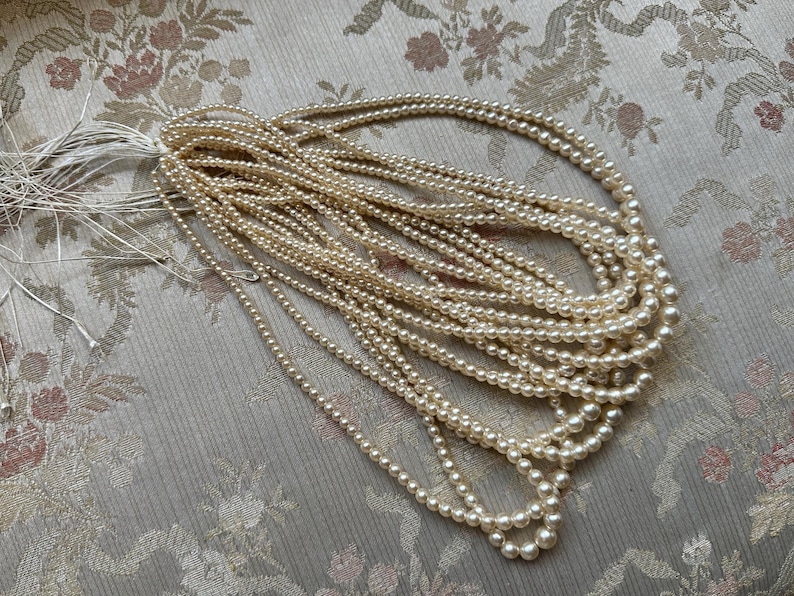 Puede incluir: Una colecci&oacute;n de collares de perlas de imitaci&oacute;n de color crema, colocados sobre una tela con estampado floral. Las perlas est&aacute;n ensartadas, creando m&uacute;ltiples hilos. Los collares son un accesorio cl&aacute;sico.