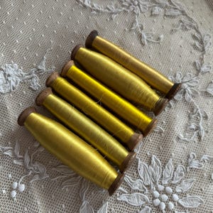 May include: Six spools of bright yellow thread are arranged diagonally on a white lace surface. The thread is wound around small wooden spools. The lace has a delicate floral pattern.