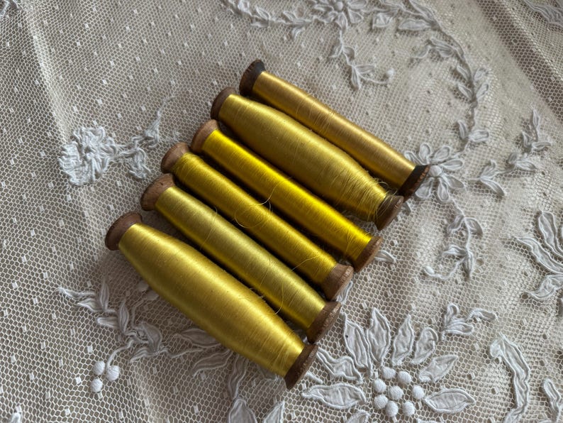 May include: Six spools of bright yellow thread are arranged diagonally on a white lace surface. The thread is wound around small wooden spools. The lace has a delicate floral pattern.