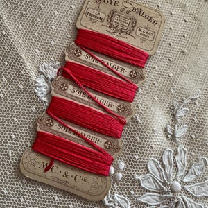 K&ouml;nnte beinhalten: Eine Sammlung roter Seidenfaden-Spulen mit der Aufschrift "Soie D'Alger" und "M.C. & Cie Paris". Die Spulen sind auf einer Karte im Vintage-Stil angeordnet, der Faden ist sauber aufgewickelt. Der Hintergrund ist eine zarte, cremefarbene Spitze.