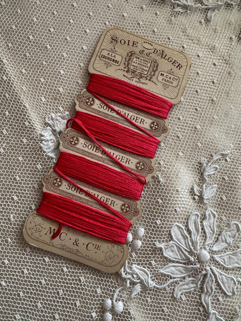 K&ouml;nnte beinhalten: Eine Sammlung roter Seidenfaden-Spulen mit der Aufschrift "Soie D'Alger" und "M.C. & Cie Paris". Die Spulen sind auf einer Karte im Vintage-Stil angeordnet, der Faden ist sauber aufgewickelt. Der Hintergrund ist eine zarte, cremefarbene Spitze.