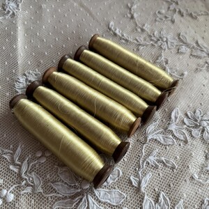 May include: Six spools of pale yellow thread are arranged diagonally. The thread is wound around small wooden spools. The background is a delicate, off-white lace fabric with floral and dotted patterns.