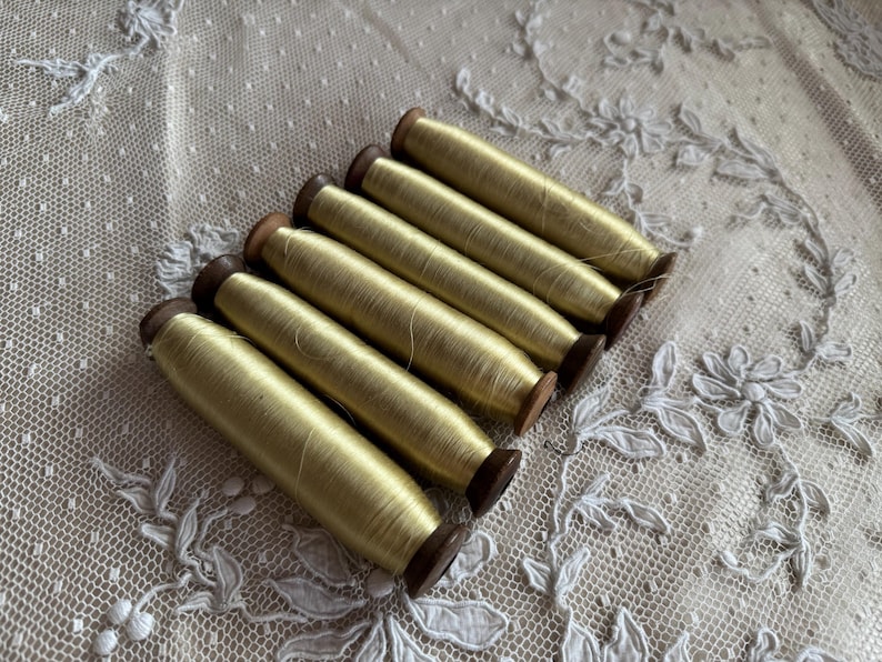 May include: Six spools of pale yellow thread are arranged diagonally. The thread is wound around small wooden spools. The background is a delicate, off-white lace fabric with floral and dotted patterns.